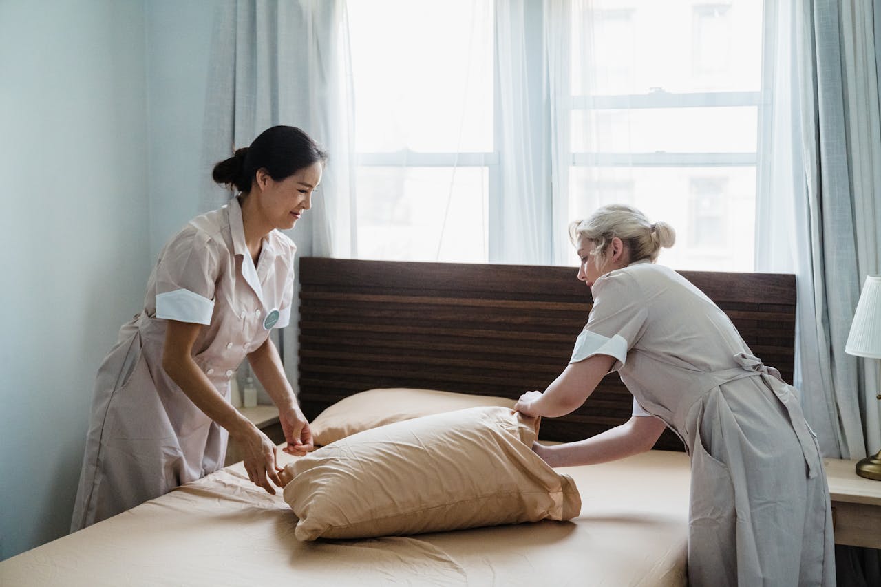 services-02 Two housekeepers in uniform collaborate while making a bed in a bright, peaceful bedroom.