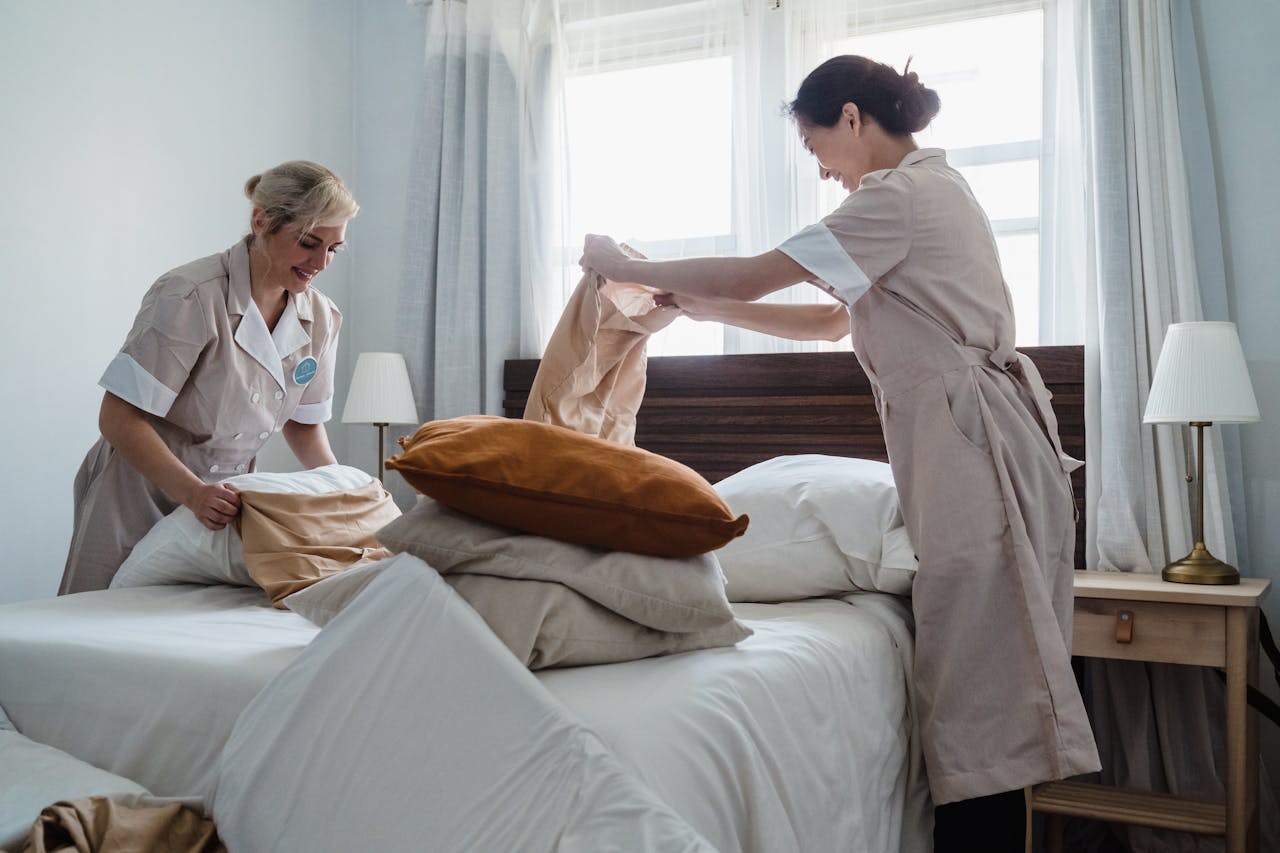hero-img-01 Two housekeepers in uniform fixing a hotel room bed with fresh linen and pillows.
